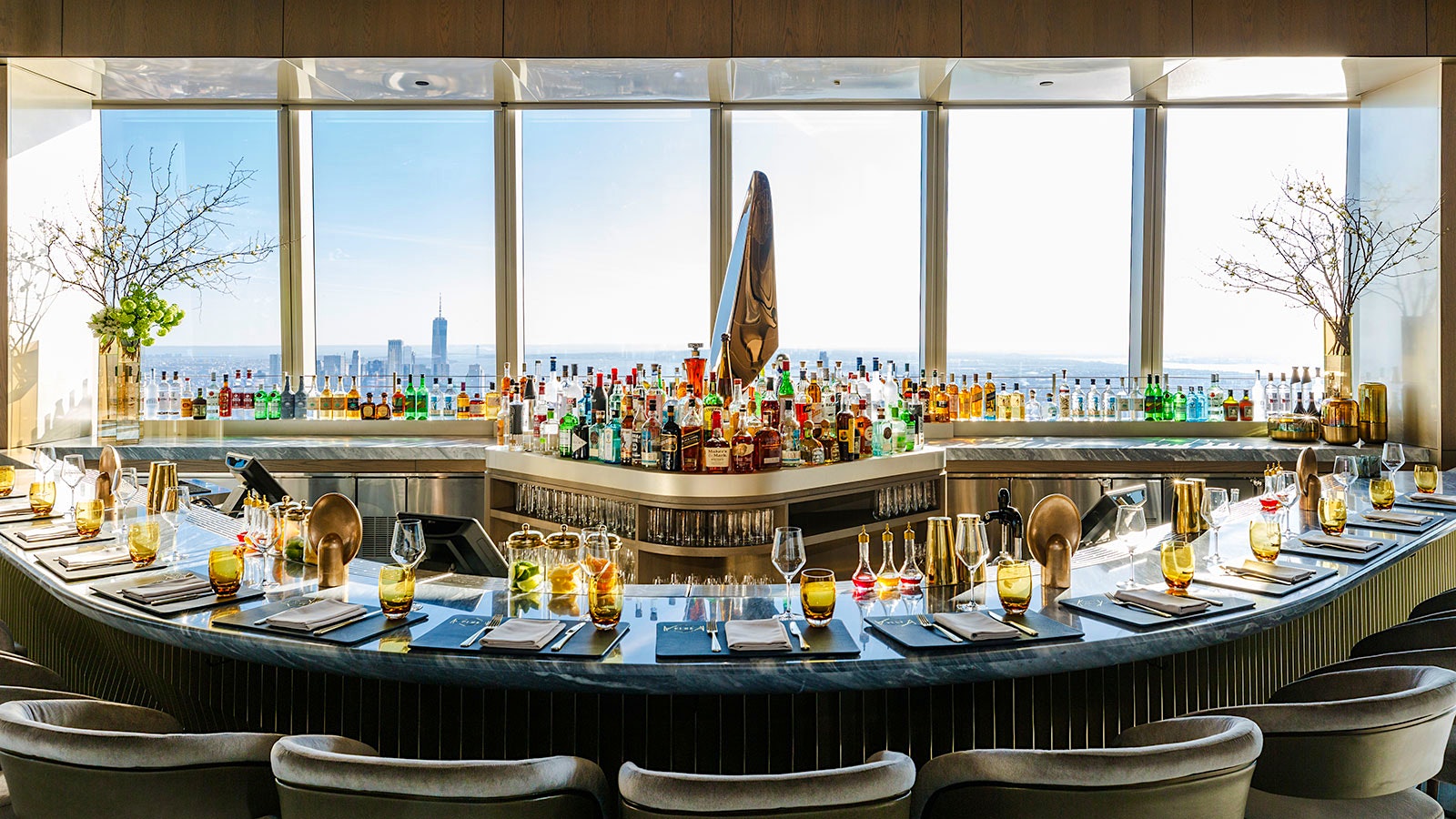 The elegant bar at Peak restaurant with a semi-circle of seats in the front of it and views over New York City behind it